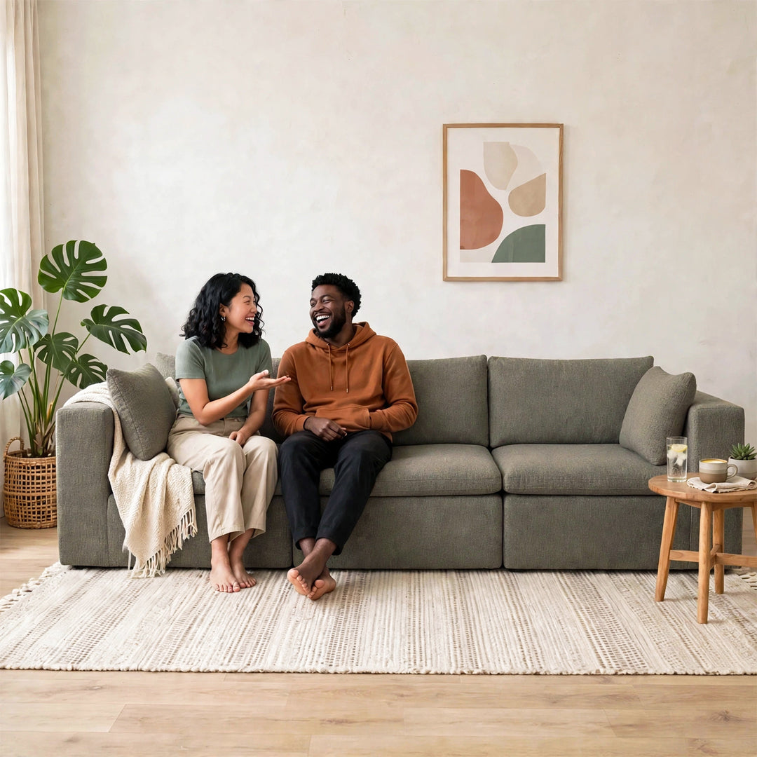 A man and a woman sit on a gray fabric sofa in a bright, modern living room. The woman wears a sage green top and cream pants, and the man wears a rust orange hoodie and dark pants. 