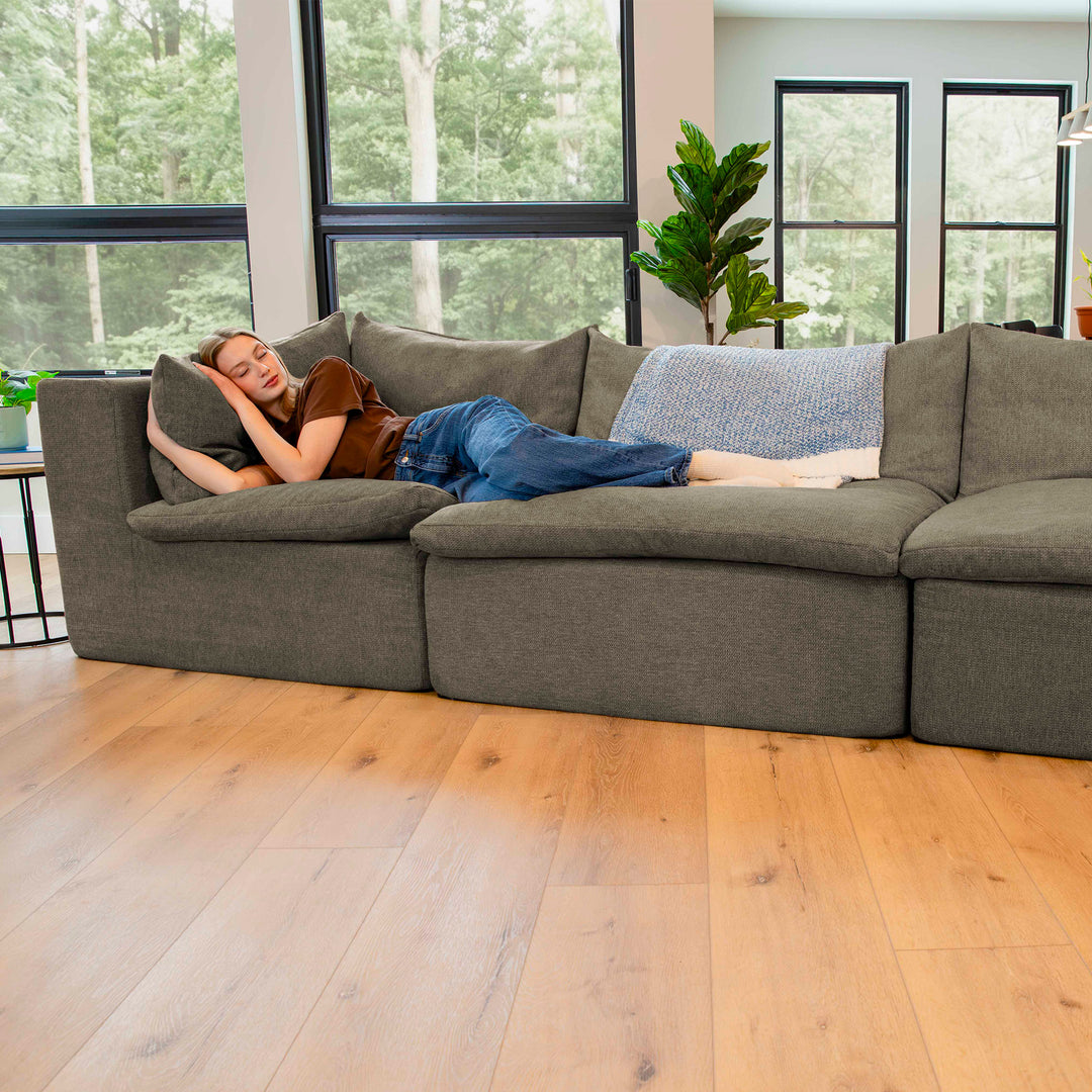 Person lying on a large gray sectional sofa in a modern living room with large windows. 