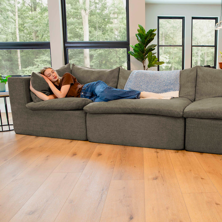 Person lying on a large gray sectional sofa in a modern living room with large windows. 