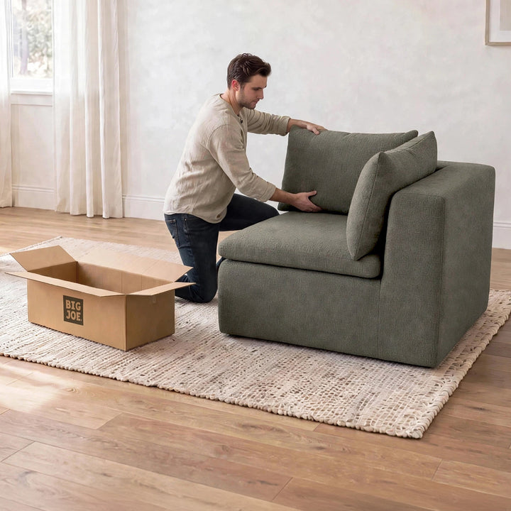 A man kneels beside a large upholstered armchair in a bright room, adjusting the back cushion. An open cardboard box labeled BIG JOE sits on the floor nearby, and the chair is placed on a woven rug over a wood floor. 