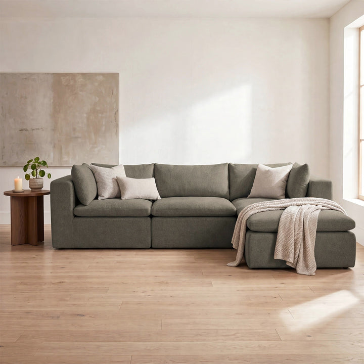 A modern sectional sofa in a muted sage green fabric, styled with neutral throw pillows and a beige knit blanket in a bright, minimalist living room with light wood floors. 