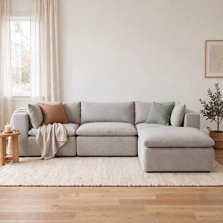 A light gray upholstered sectional sofa with a right-facing chaise ottoman, styled in a bright, modern living room with neutral-toned pillows, a throw blanket, a small wooden side table, and a potted plant. 