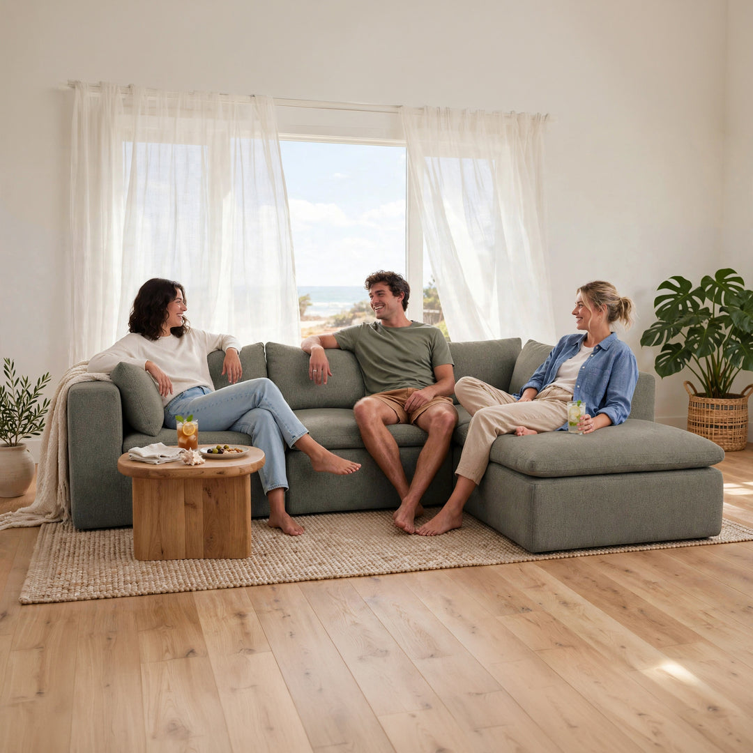 A three-person sofa in a bright, modern living room with a wooden coffee table, a woven rug, and large windows with sheer white curtains. Two people are sitting on the sofa, and a third person is seated on the chaise lounge, with a potted plant on the right. 
