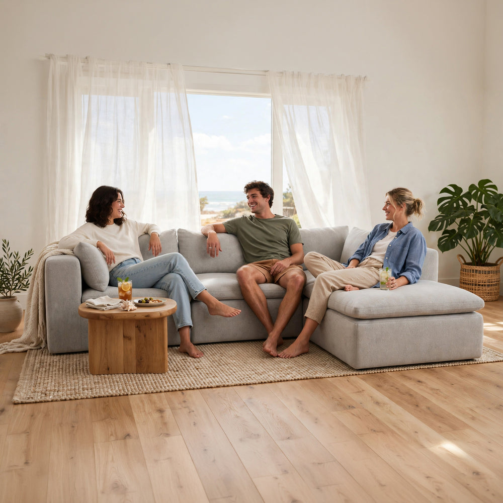 A three-person sofa in a bright, modern living room with a wooden coffee table, a large window with sheer white curtains, and potted plants. Two people are sitting on the sofa, and a woman in a blue denim jacket sits on the chaise ottoman. #color_cottage-gray-terra-knit