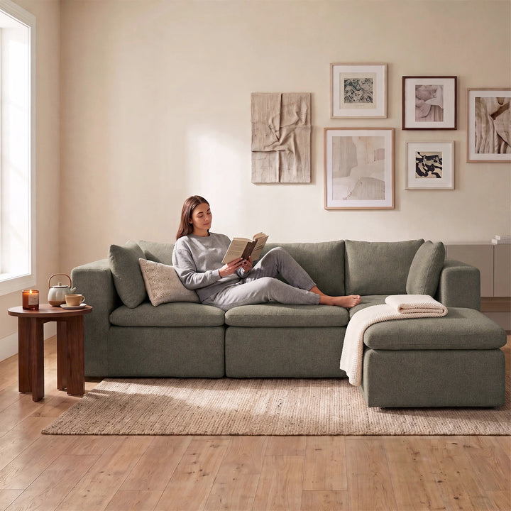 A woman in a gray lounge set sits on a large sage green sectional sofa in a bright, modern living room, reading a book. The sofa features a chaise lounge on the right side with a folded cream throw blanket, and the room has a light wood floor, a beige rug, and framed wall art. 