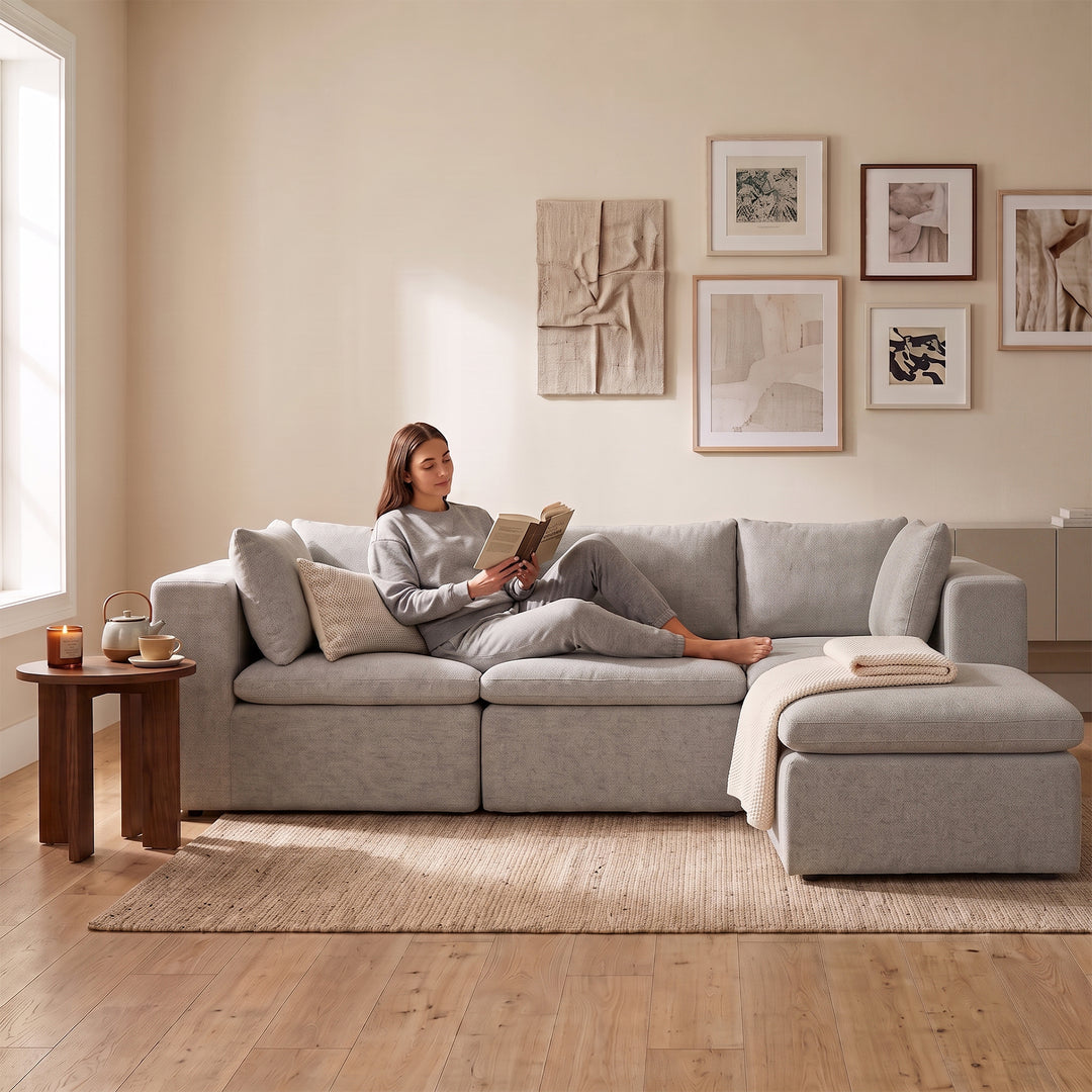 A woman in a light gray lounge set sits on a light gray sectional sofa with a chaise in a bright, modern living room. The room features a beige wall with framed abstract art, a wooden side table with a teapot, and a light wood floor. 