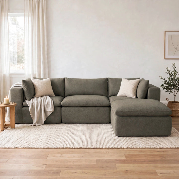 A large L-shaped sectional sofa in a muted sage green fabric, styled in a bright, modern living room with a cream woven rug, light wood floor, and neutral throw pillows. 