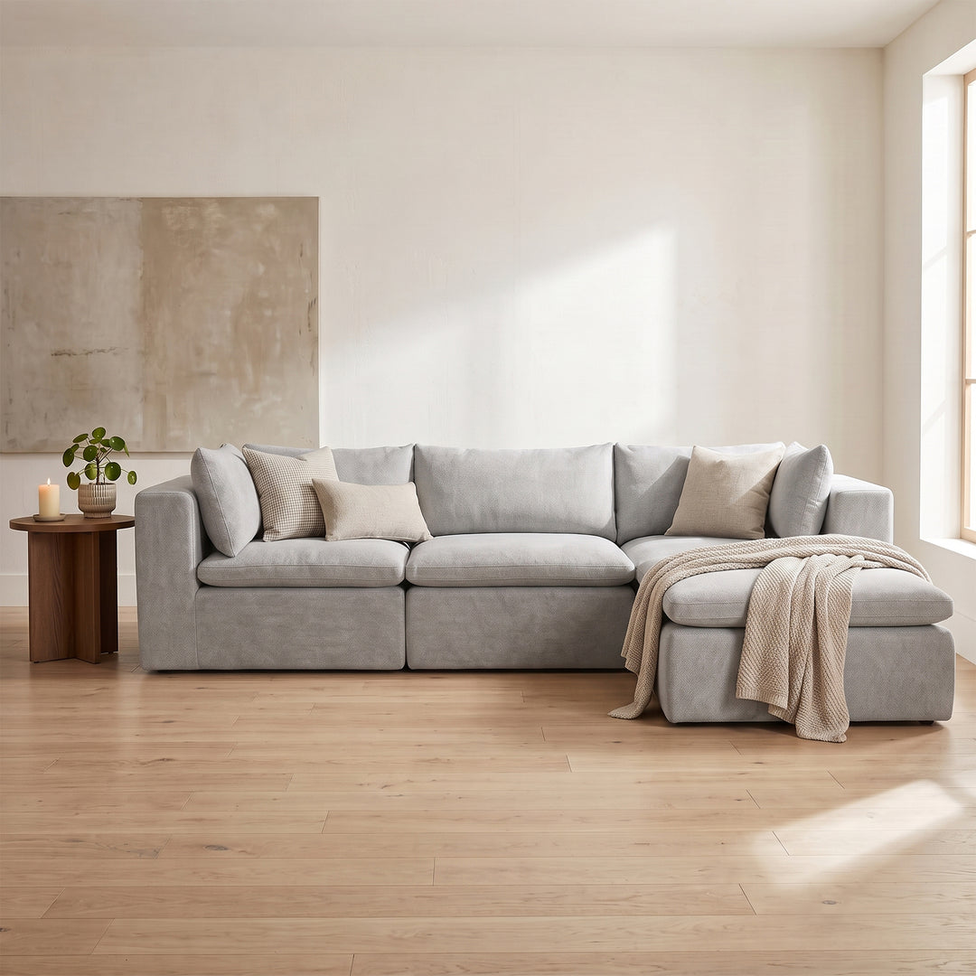 A light gray upholstered sectional sofa with a chaise lounge, styled with neutral beige throw pillows and a beige throw blanket in a bright, modern living room with light wood floors. #color_cottage-gray-terra-knit