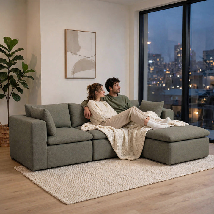 A man and a woman relax on a large, modern gray sectional sofa in a cozy living room with a city view at night, styled with a cream knit blanket and a textured rug. 
