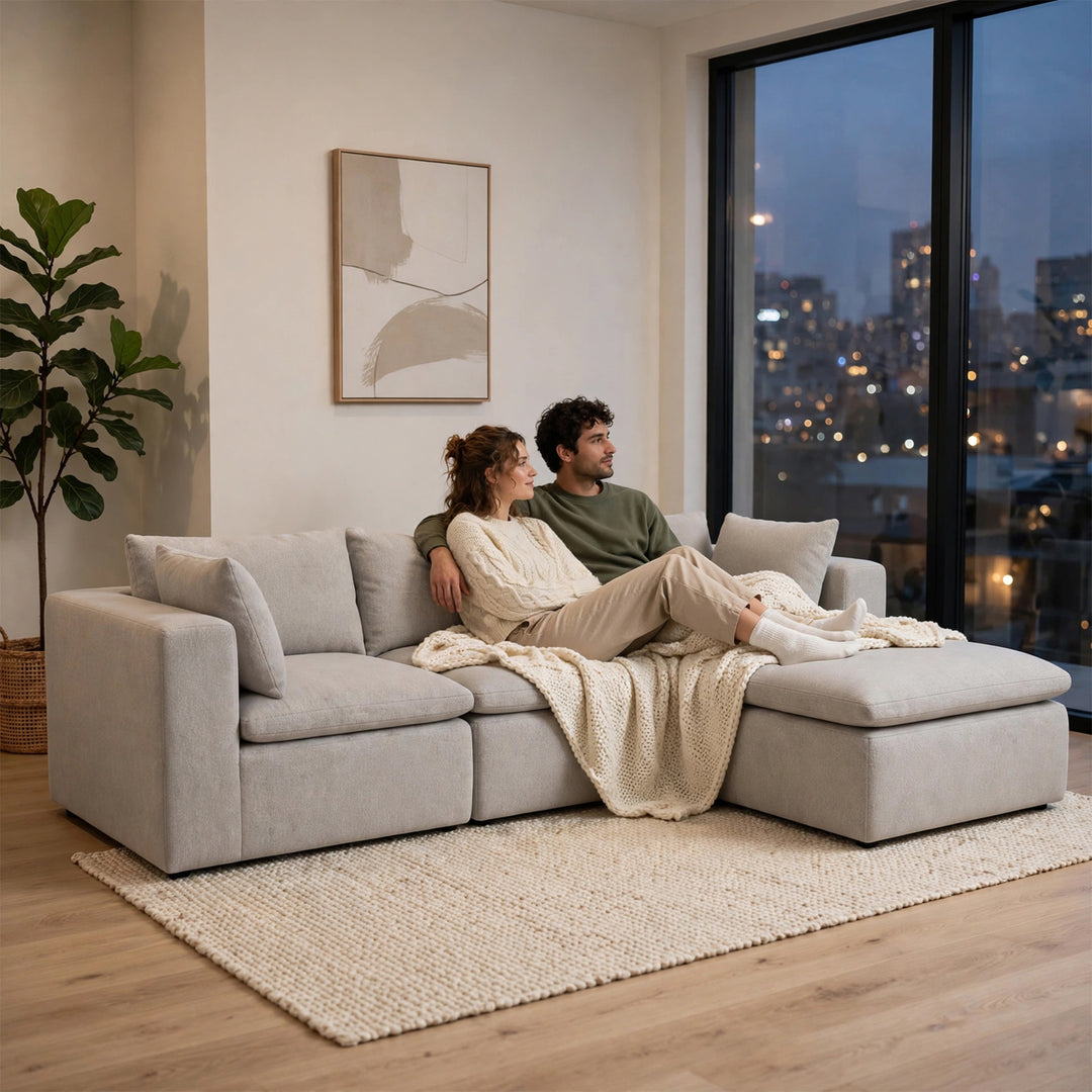 A couple relaxes on a light gray modular sectional sofa in a modern living room, with a woman leaning on a man and a cream knit throw blanket draped across the seat. The room features a large window showing a city skyline at dusk, a potted plant, and a textured rug. 