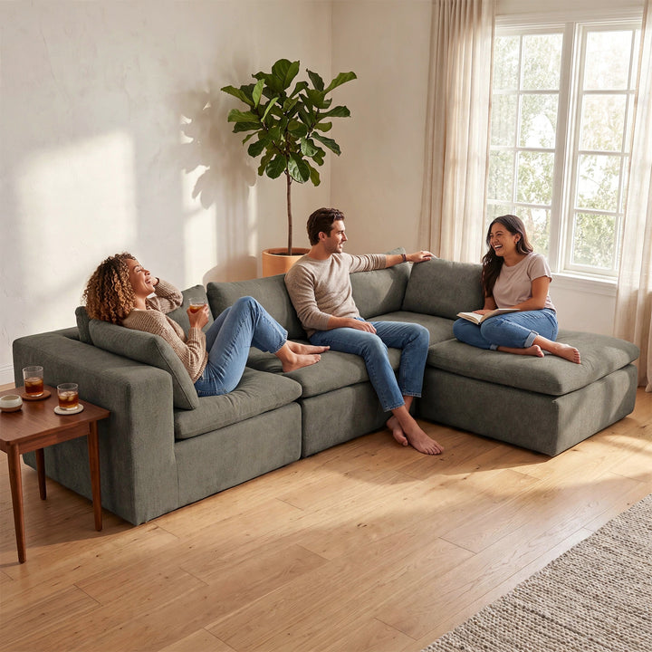 A three-person sectional sofa in a sage green fabric, shown in a bright, modern living room with two people sitting on it and a third person reclining. A small wooden side table with drinks is on the left, and a large potted fiddle-leaf fig plant stands near the window in the background. 