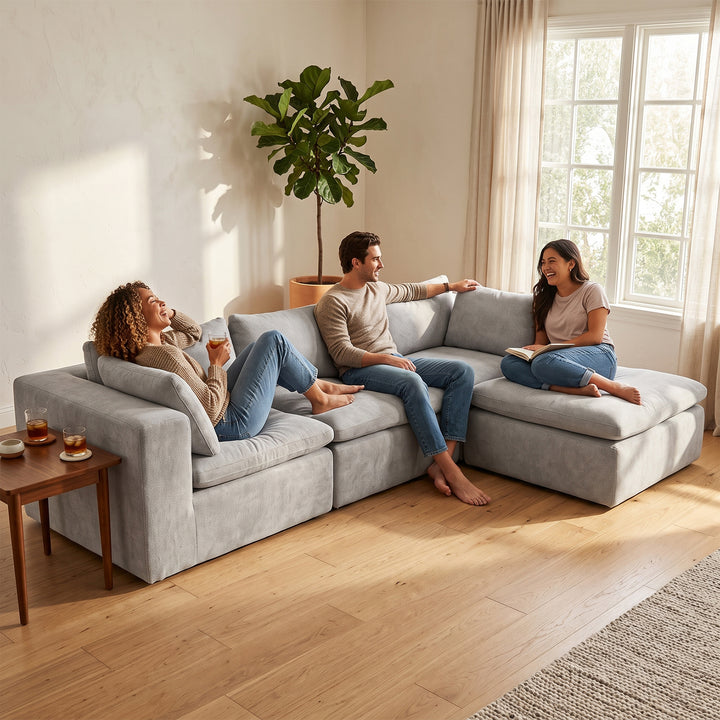 A light gray modular sectional sofa in a bright, modern living room with three people sitting and relaxing on it. The room features light wood floors, beige walls, a large potted plant, and a window with sheer curtains. 