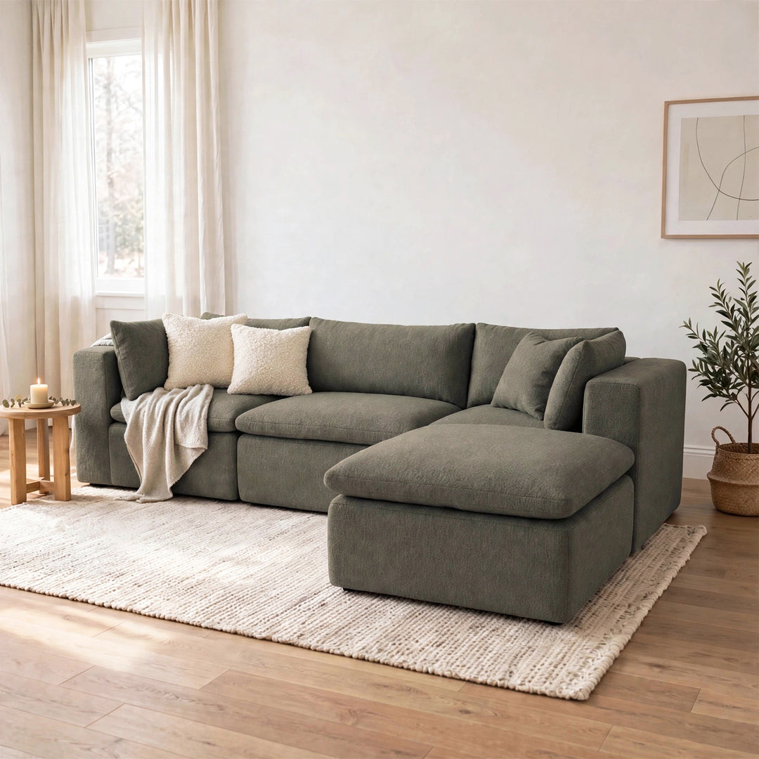 A modern sectional sofa with a chaise lounge in a muted sage green fabric, styled in a bright, minimalist living room with a cream rug, wooden floor, and a small side table. 