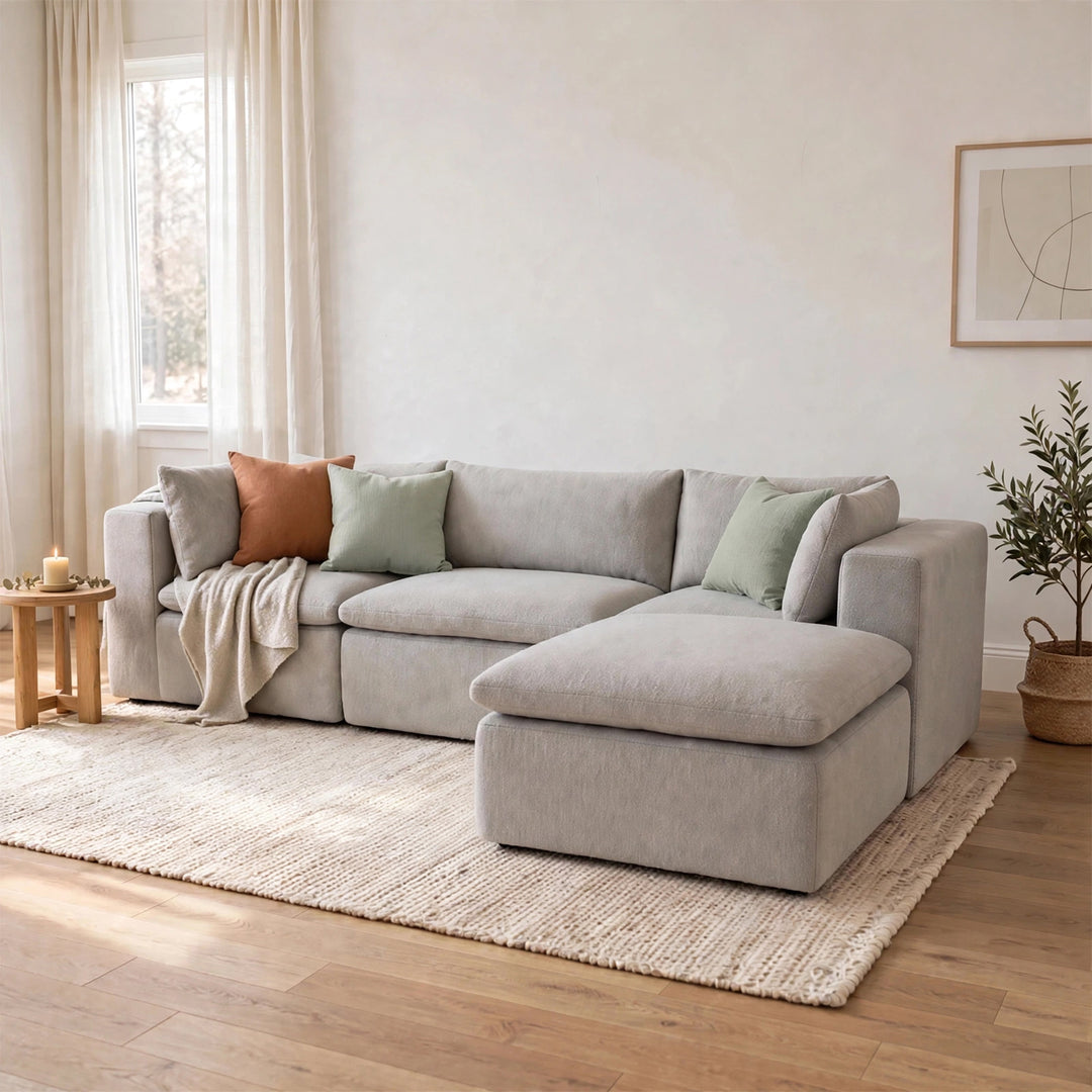 A light gray upholstered sectional sofa with a chaise lounge, styled in a bright, modern living room with neutral-toned walls, a woven rug, and decorative pillows. 
