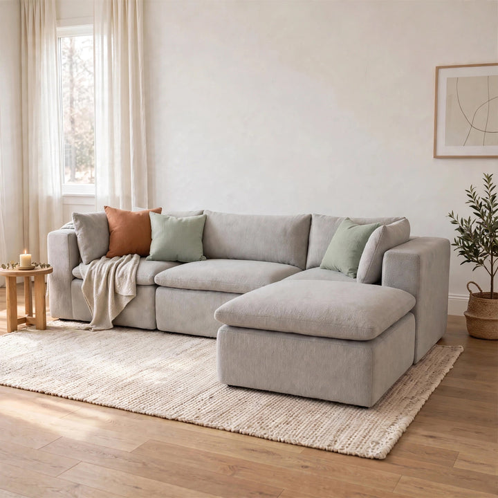 A light gray upholstered sectional sofa with a chaise lounge, styled in a bright, modern living room with neutral-toned walls, a woven rug, and decorative pillows. 