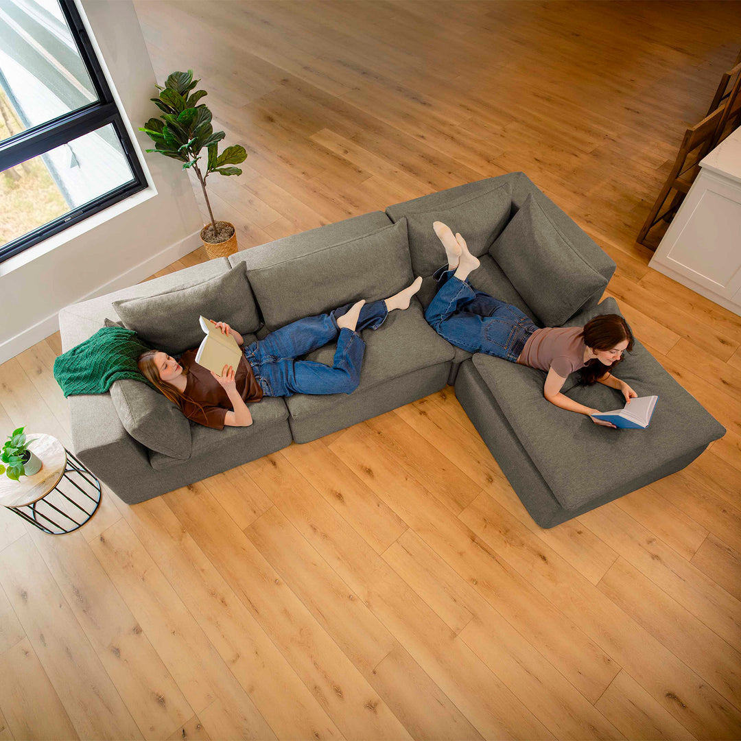 Two people lying on a gray sectional sofa in a living room with wooden floors. 