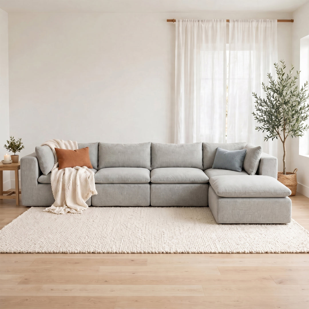 A light gray modular sectional sofa with a chaise lounge, styled in a bright, modern living room with a cream rug, wooden floor, and sheer white curtains. #color_cottage-gray-terra-knit