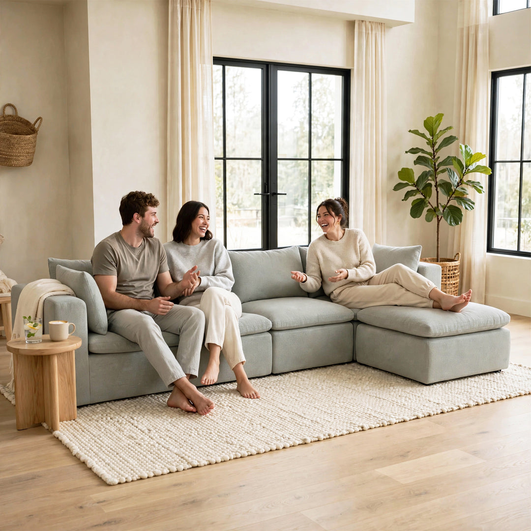 A light gray modular sectional sofa in a bright, modern living room with three people sitting and relaxing on it. The room features beige walls, large black-framed windows, a large white textured area rug, and a potted fiddle-leaf fig plant. 