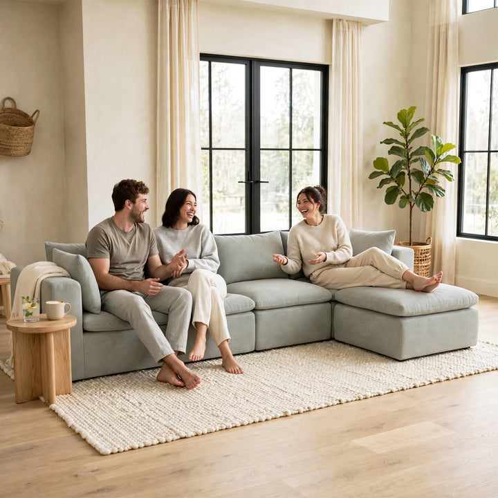 A light gray modular sectional sofa in a bright, modern living room with three people sitting and relaxing on it. The room features beige walls, large black-framed windows, a large white textured area rug, and a potted fiddle-leaf fig plant. 