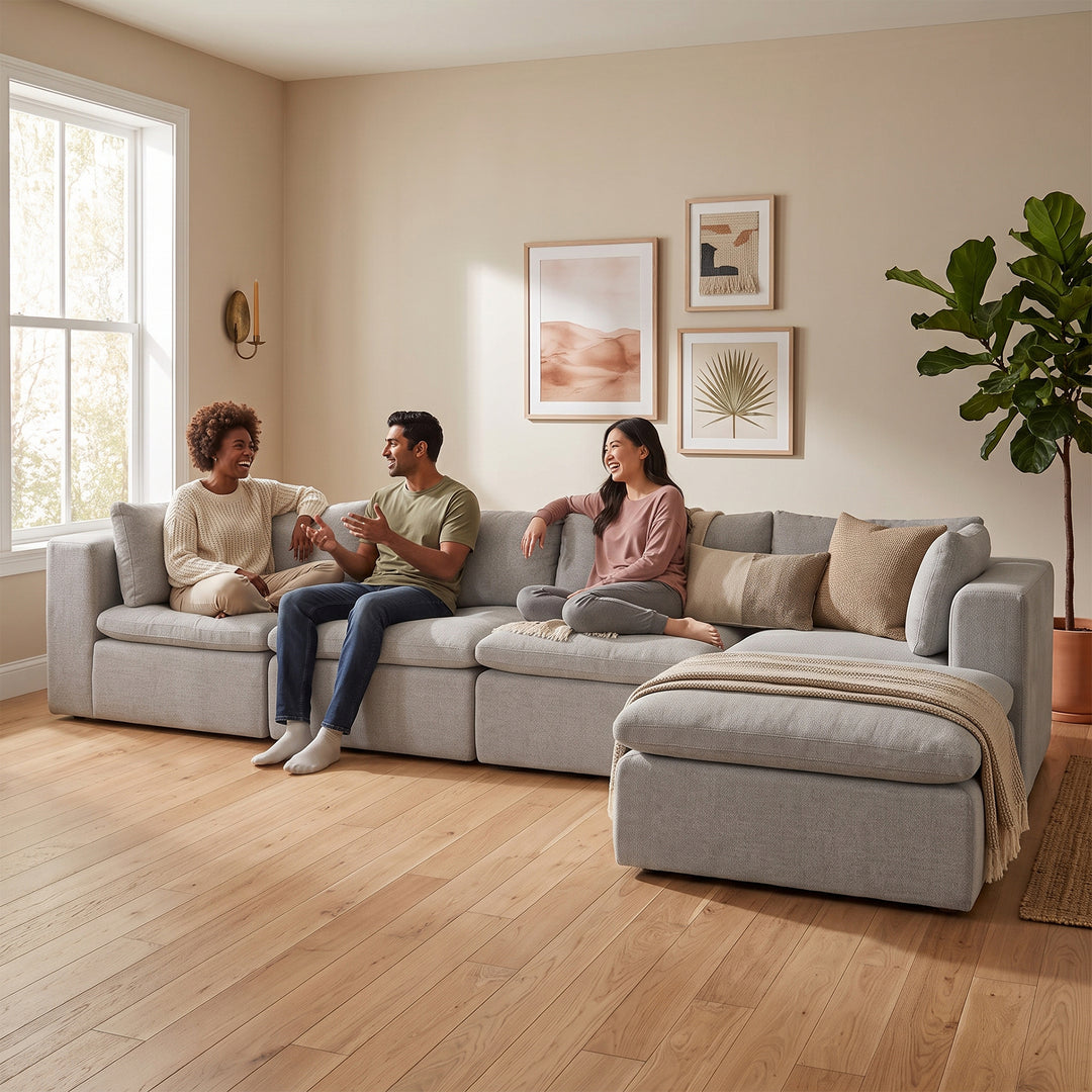 A light gray sectional sofa with a chaise lounge is shown in a bright, modern living room with three people sitting on it. The room has beige walls, light wood floors, framed wall art, and a potted plant. 