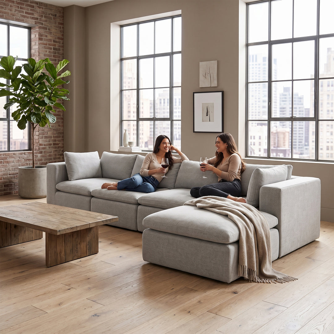 A light gray modular sectional sofa with two women sitting on it in a bright, modern living room with large windows, a wooden coffee table, and a potted plant. 