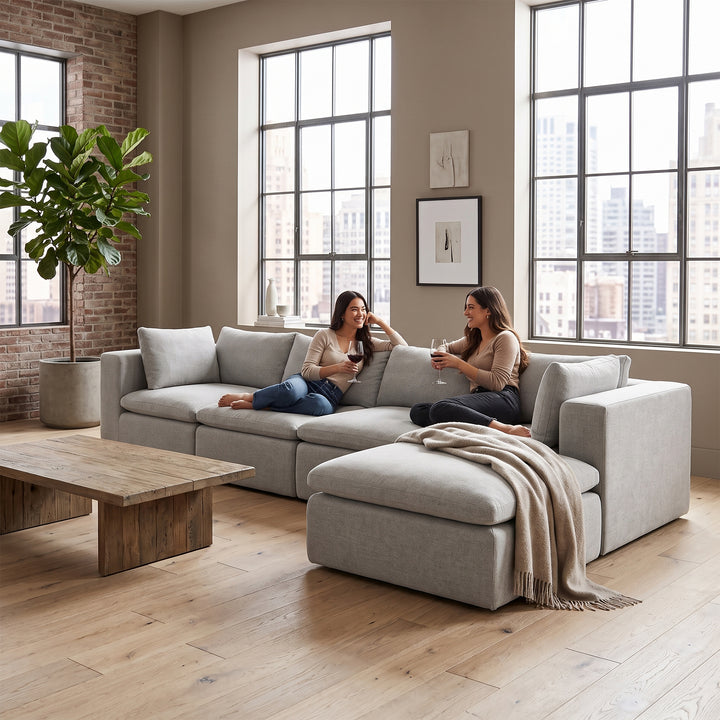 A light gray modular sectional sofa with two women sitting on it in a bright, modern living room with large windows, a wooden coffee table, and a potted plant. 