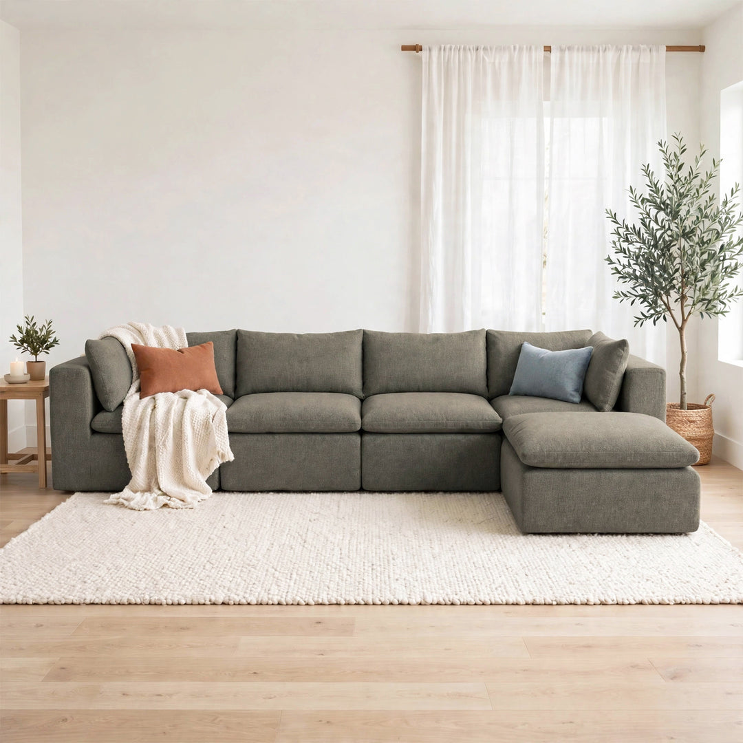 A modern L-shaped sectional sofa upholstered in textured gray fabric, styled in a bright, minimalist living room with a cream shag rug, light wood floor, and white curtains. 