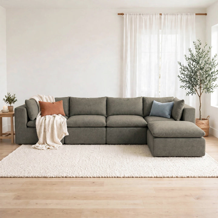 A modern L-shaped sectional sofa upholstered in textured gray fabric, styled in a bright, minimalist living room with a cream shag rug, light wood floor, and white curtains. 
