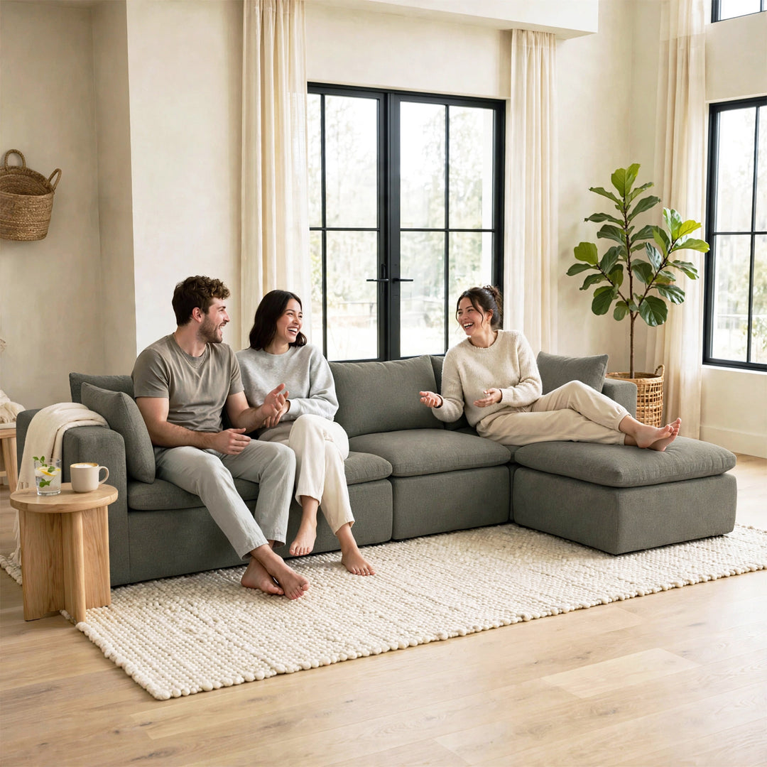 Three people relax on a large gray sectional sofa in a bright, modern living room with beige walls, black-framed windows, a white textured rug, and a potted plant. 