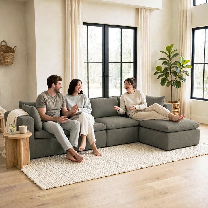 Three people relax on a large gray sectional sofa in a bright, modern living room with beige walls, black-framed windows, a white textured rug, and a potted plant. 