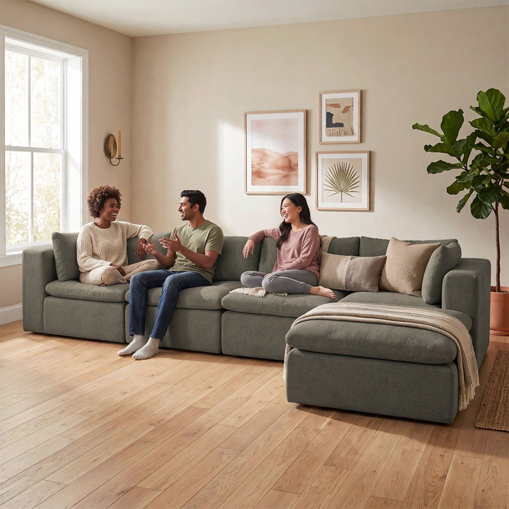 A three-person sectional sofa in a sage green fabric, styled with neutral throw pillows and a beige throw blanket, with three people sitting on it in a bright, modern living room with light wood floors. 