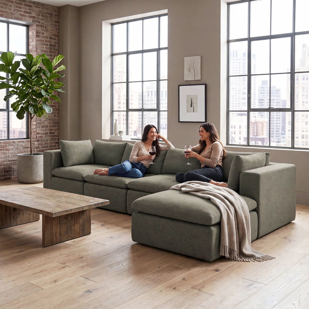 A large sage green sectional sofa with a chaise lounge in a modern loft-style living room, featuring two women sitting on the couch and a wooden coffee table in the foreground. 