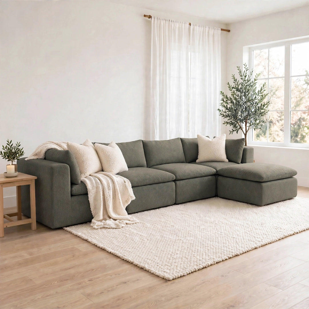 A modern sectional sofa in a muted sage green color, styled with cream throw pillows and a cream knit blanket in a bright, minimalist living room with light wood floors and a large white rug. 