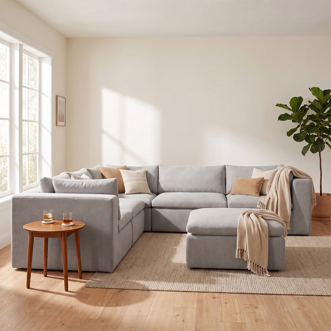 A light gray sectional sofa with a matching ottoman in a bright, modern living room. The sofa is styled with neutral beige and tan pillows and a beige throw blanket, set on a woven jute rug next to a small wooden side table and a potted plant. 