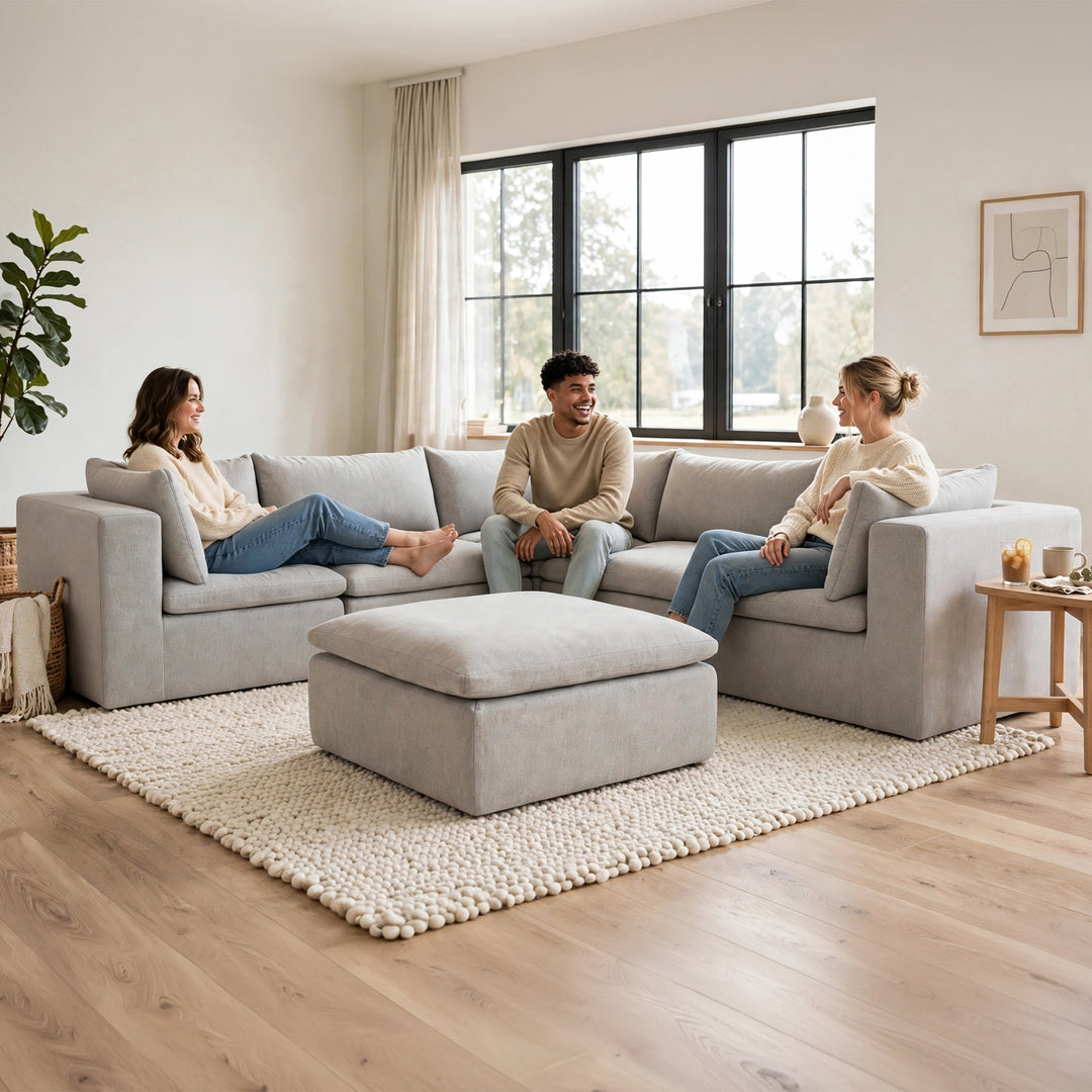 A light gray modular sectional sofa with a matching ottoman in a bright, modern living room. Two people are sitting on the sofa, and a third person is visible on the right side. 