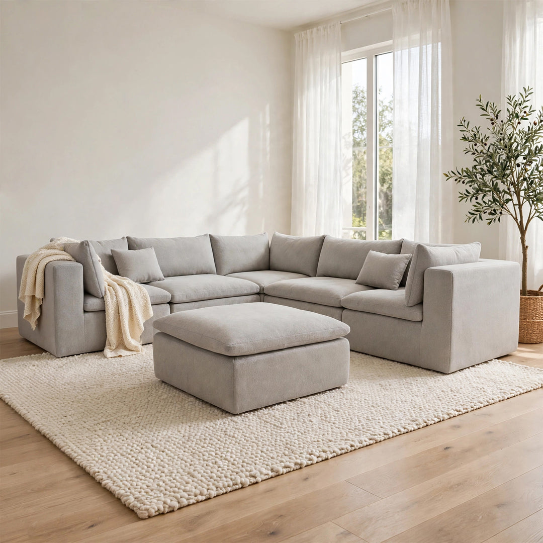 A light gray upholstered sectional sofa with matching throw pillows and a matching ottoman, staged in a bright, modern living room with a textured cream rug, wood floor, sheer curtains, and a potted plant. 