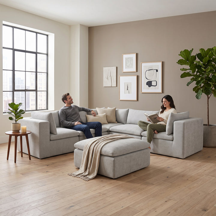 A modern light gray fabric sectional sofa with an ottoman in a bright, contemporary living room. A man sits on the left side of the sofa, and a woman sits on the right side, reading a book. 