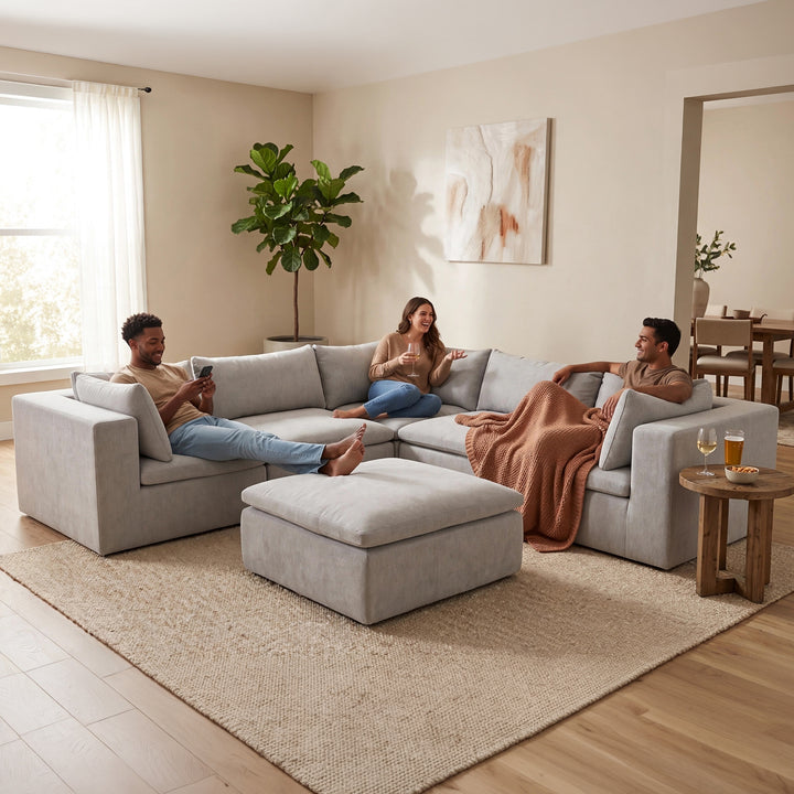 A light gray fabric sectional sofa with an ottoman in a bright, modern living room. Three people are lounging on the sofa and ottoman, with one person draped a brown blanket over their lap. 