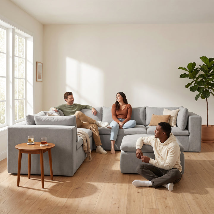 A modern light gray sectional sofa with matching armchair and ottoman in a bright, sunlit living room, with two people sitting on the furniture and a third person sitting on the floor. 