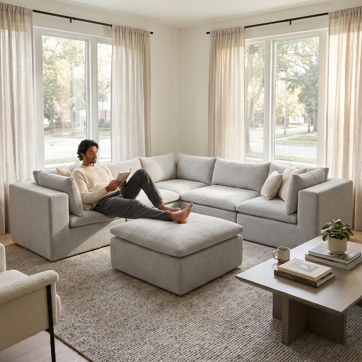 A man relaxes on a light gray sectional sofa in a bright, modern living room with large windows, beige curtains, and a matching ottoman. 
