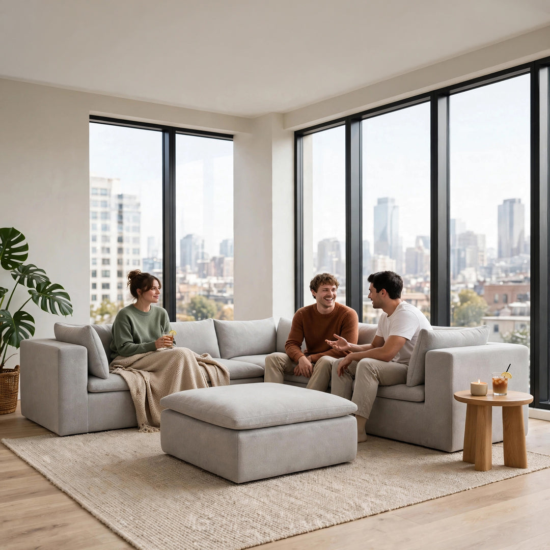 A light gray modular sectional sofa with a matching ottoman in a bright, modern living room. Three people are sitting on the sofa, and large windows with a city skyline view are in the background. 