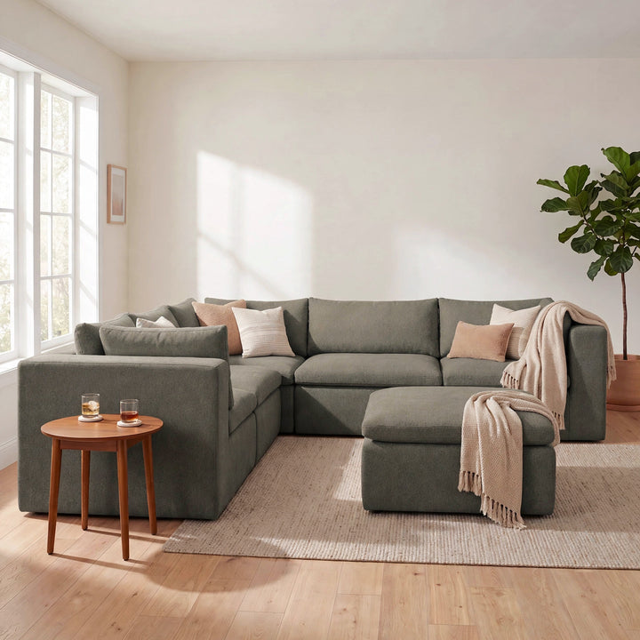 A modern L-shaped sectional sofa in a muted sage green fabric, styled with neutral throw pillows and a beige throw blanket in a bright, sunlit living room. 
