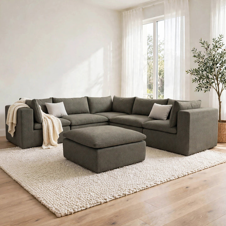 A modern L-shaped sectional sofa in a dark gray fabric, paired with a matching square ottoman, styled in a bright, minimalist living room with a cream shag rug, light wood floors, and a potted tree.  