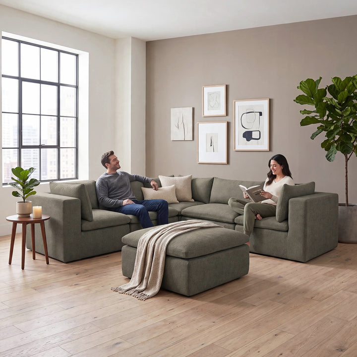 A modern sectional sofa set in a bright, contemporary living room with a man and woman sitting on the couch, with a matching ottoman in front. The room features light wood floors, a beige wall with minimalist framed art, and large windows.  