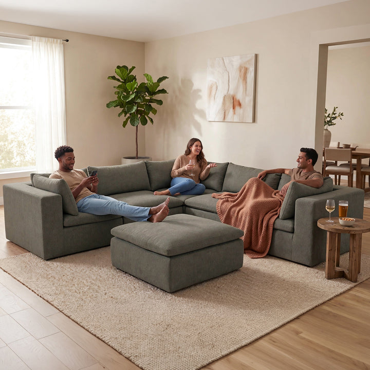 A three-person gray fabric sectional sofa with an ottoman in a modern, warm-toned living room. Two men and a woman are lounging on the sofa, with one man holding a phone and another draped with a brown throw blanket. 