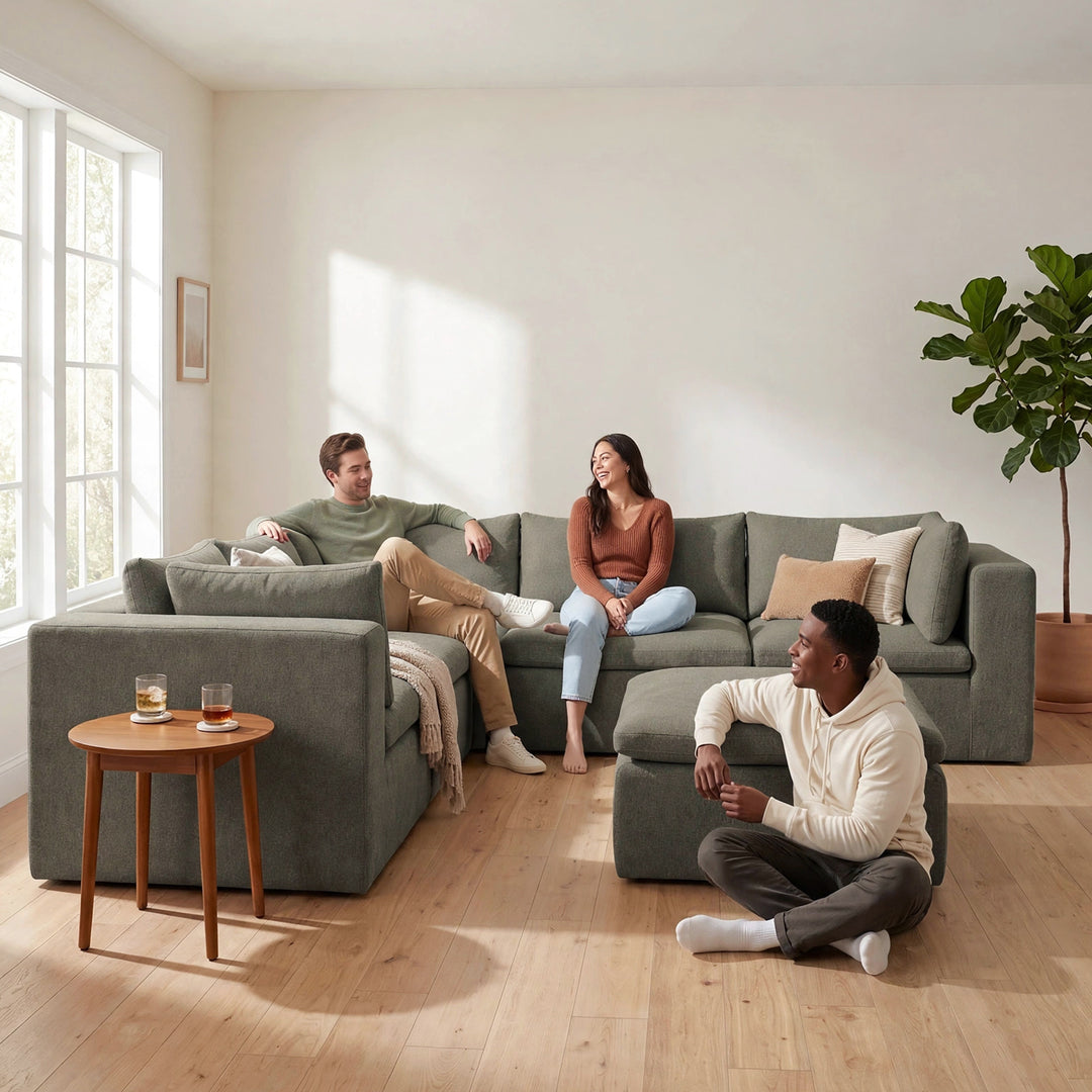 A three-person gray sectional sofa in a bright, modern living room with light wood floors, a woman sitting on the sofa, a man sitting on the left armchair, and another man sitting on the floor in front of the sofa. 