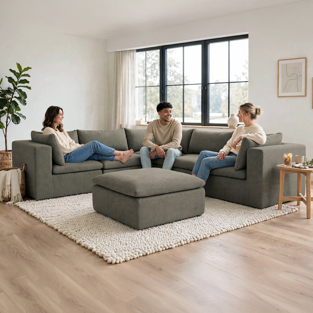 A three-person gray fabric sectional sofa with a matching ottoman in a bright, modern living room. Two women and one man are sitting on the sofa, which is placed on a chunky white shag rug over light wood flooring. 