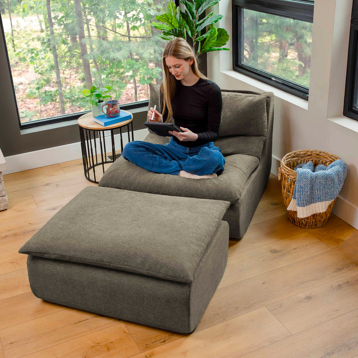 Woman sitting on a gray sectional sofa in a room with large windows and a plant. 