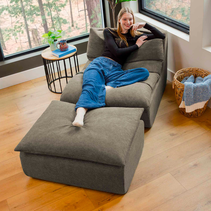 Woman sitting on a gray sectional sofa in a room with large windows and wooden flooring. 
