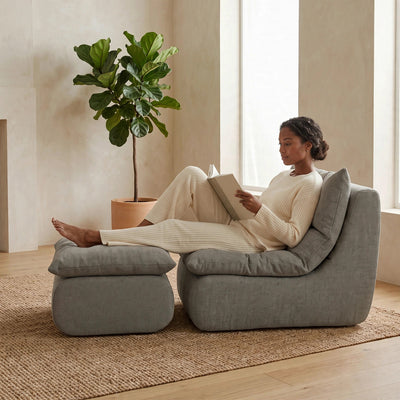 A woman in cream loungewear reclines on a gray upholstered lounge chair with an ottoman in a bright, neutral room, holding an open book. #color_misty-gray-terra-knit